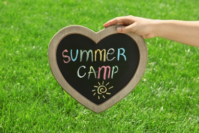 Photo of Woman holding heart shaped blackboard with text SUMMER CAMP and drawing over green grass