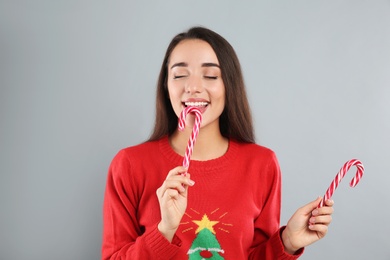 Young woman in Christmas sweater holding candy canes on grey background