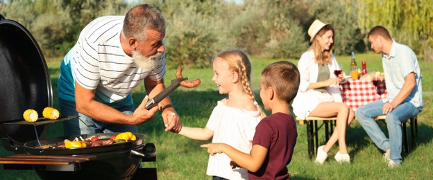 Image of Grandfather with little kids cooking food on barbecue grill and their family in park. Banner design
