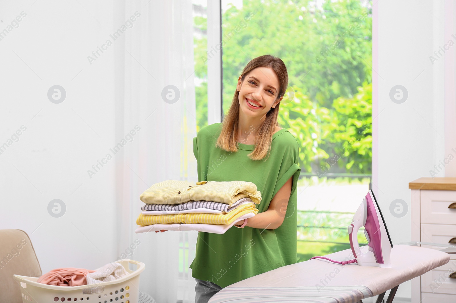 Photo of Young pretty woman holding stack of clean laundry near ironing board indoors