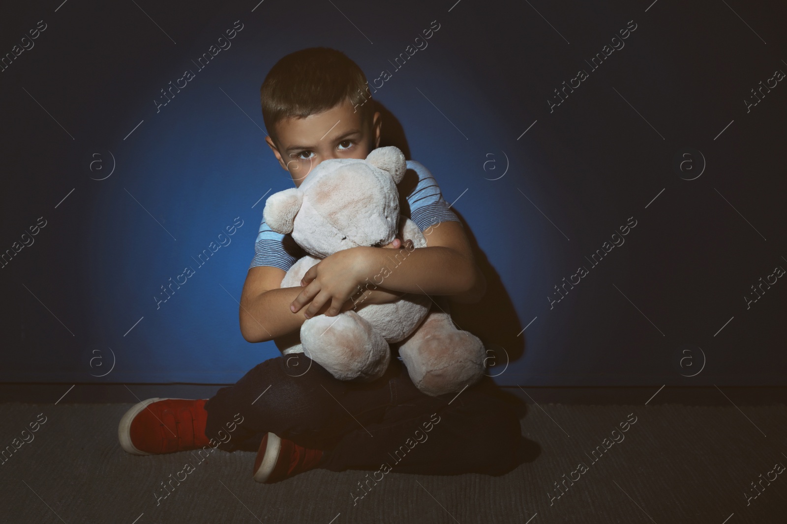 Photo of Sad little boy with teddy bear near blue wall. Domestic violence concept