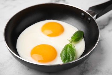 Photo of Tasty fried eggs with basil in pan on white marble table, closeup