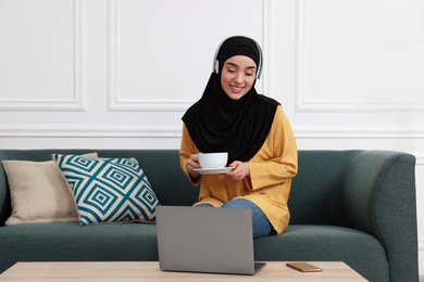 Photo of Muslim woman in hijab with cup of coffee using laptop on sofa indoors. Space for text