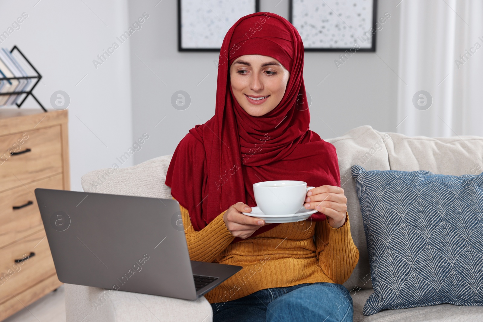 Photo of Muslim woman with cup of drink using laptop at couch in room