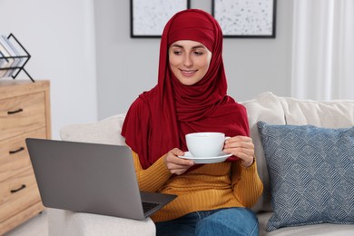 Muslim woman with cup of drink using laptop at couch in room