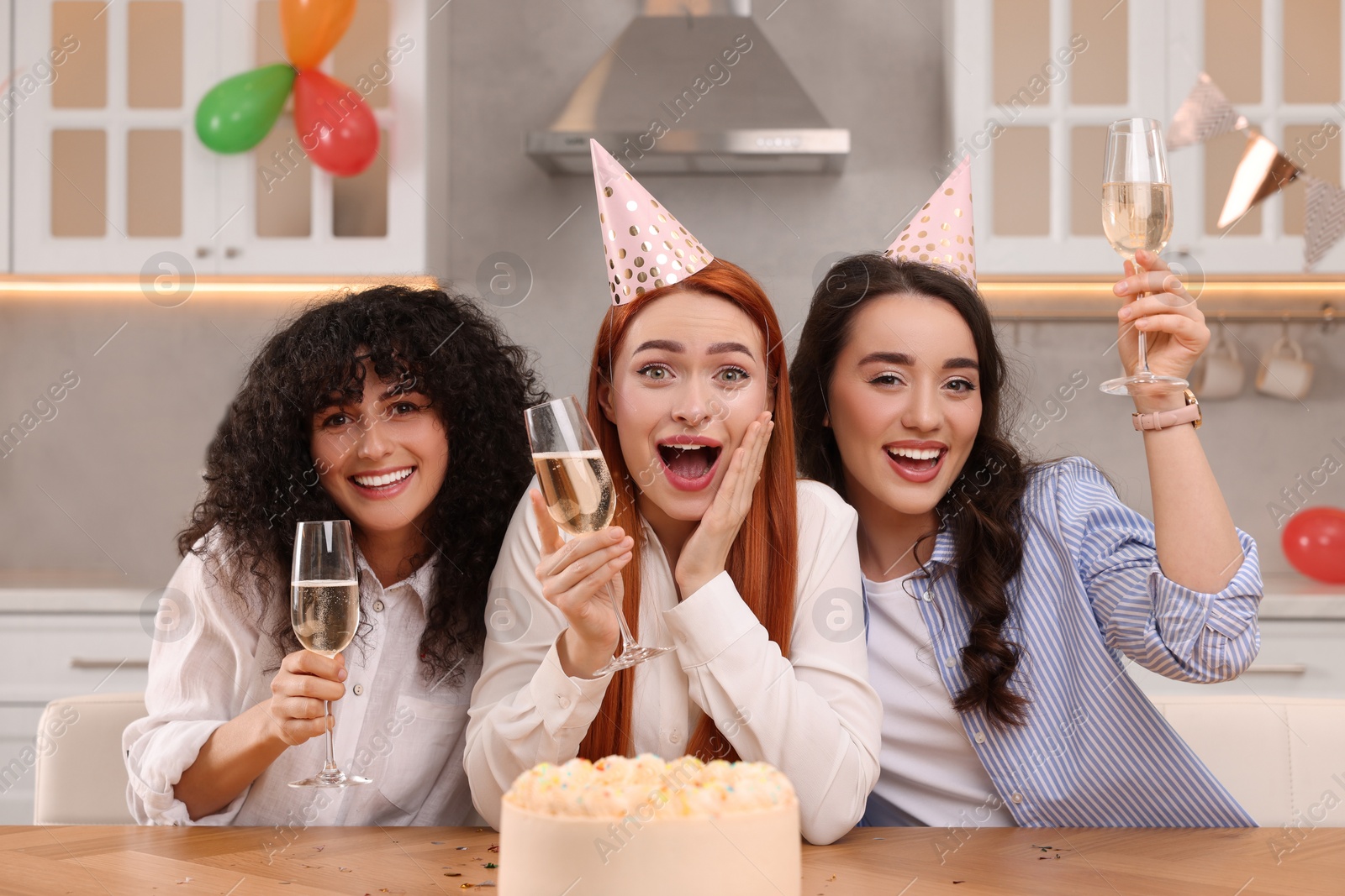 Photo of Happy young women with tasty cake and glasses of sparkling wine celebrating birthday in kitchen