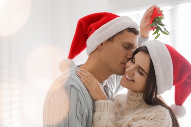 Photo of Happy man kissing his girlfriend under mistletoe bunch at home, bokeh effect
