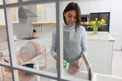 Happy young woman washing plate above sink in modern kitchen