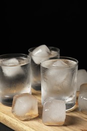 Photo of Shot glasses of vodka with ice cubes on wooden board against black background, closeup