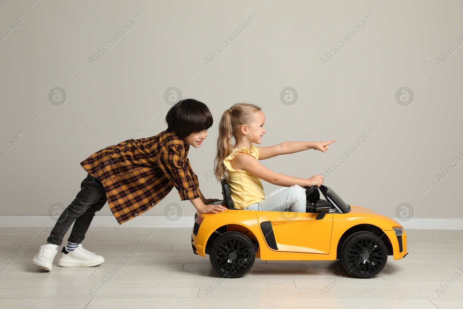 Photo of Cute boy pushing children's electric toy car with little girl near grey wall indoors