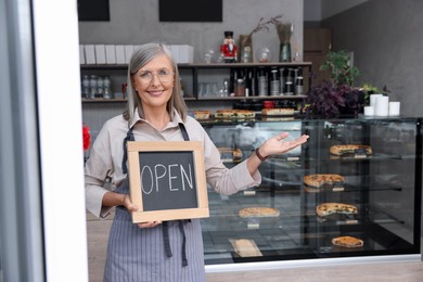 Photo of Happy business owner holding open sign and inviting to come into her cafe, space for text