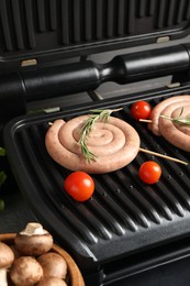 Photo of Electric grill with homemade sausages, rosemary, tomatoes and mushrooms on table