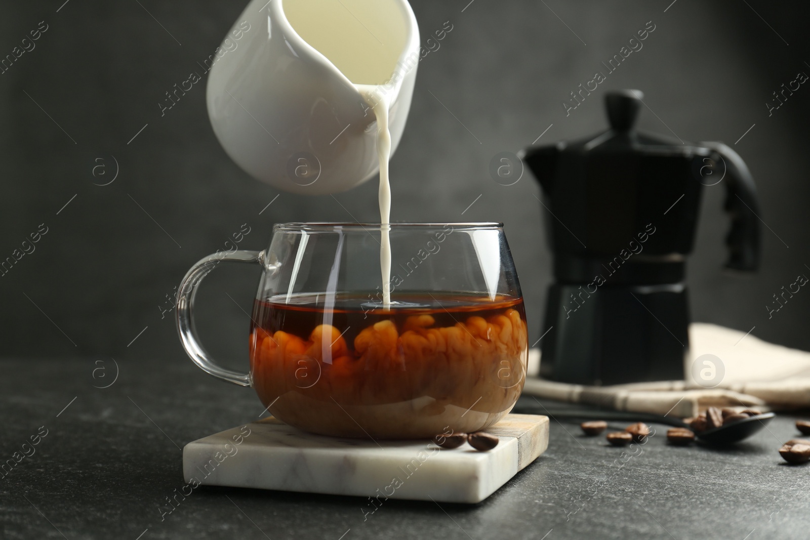 Photo of Pouring milk from pitcher into glass cup with coffee at dark textured table, closeup