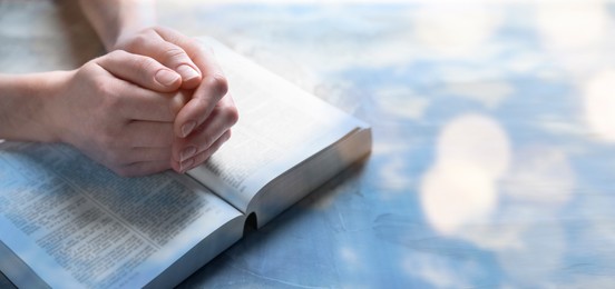 Image of Religion. Christian woman praying over Bible at table, closeup. Bokeh effect. Banner design