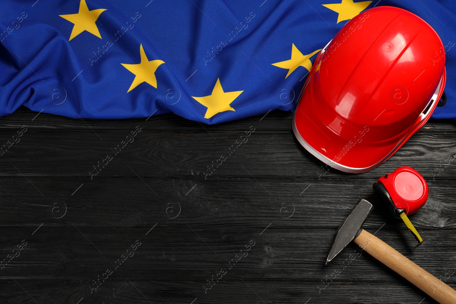 Photo of Red hard hat, measuring tape, hammer and flag of European Union on wooden black table, flat lay. Space for text