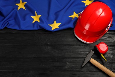 Red hard hat, measuring tape, hammer and flag of European Union on wooden black table, flat lay. Space for text