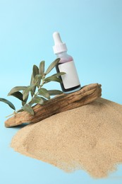Photo of Bottle with serum, olive twig and bark on sand against light blue background. Cosmetic product