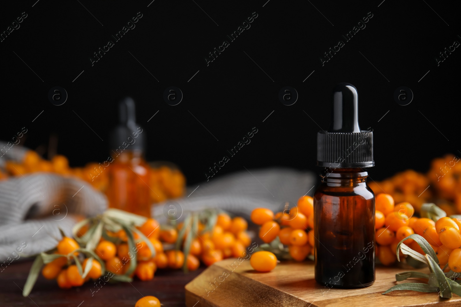 Photo of Ripe sea buckthorn and bottle of essential oil on wooden board against black background. Space for text