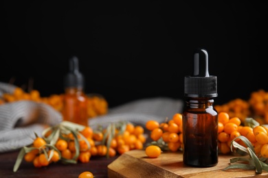 Photo of Ripe sea buckthorn and bottle of essential oil on wooden board against black background. Space for text