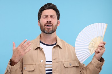 Photo of Unhappy man with hand fan suffering from heat on light blue background