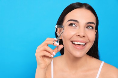 Beautiful young woman using eyelash curler on light blue background, space for text