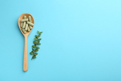 Photo of Vitamin capsules in spoon and green branch on light blue background, flat lay. Space for text