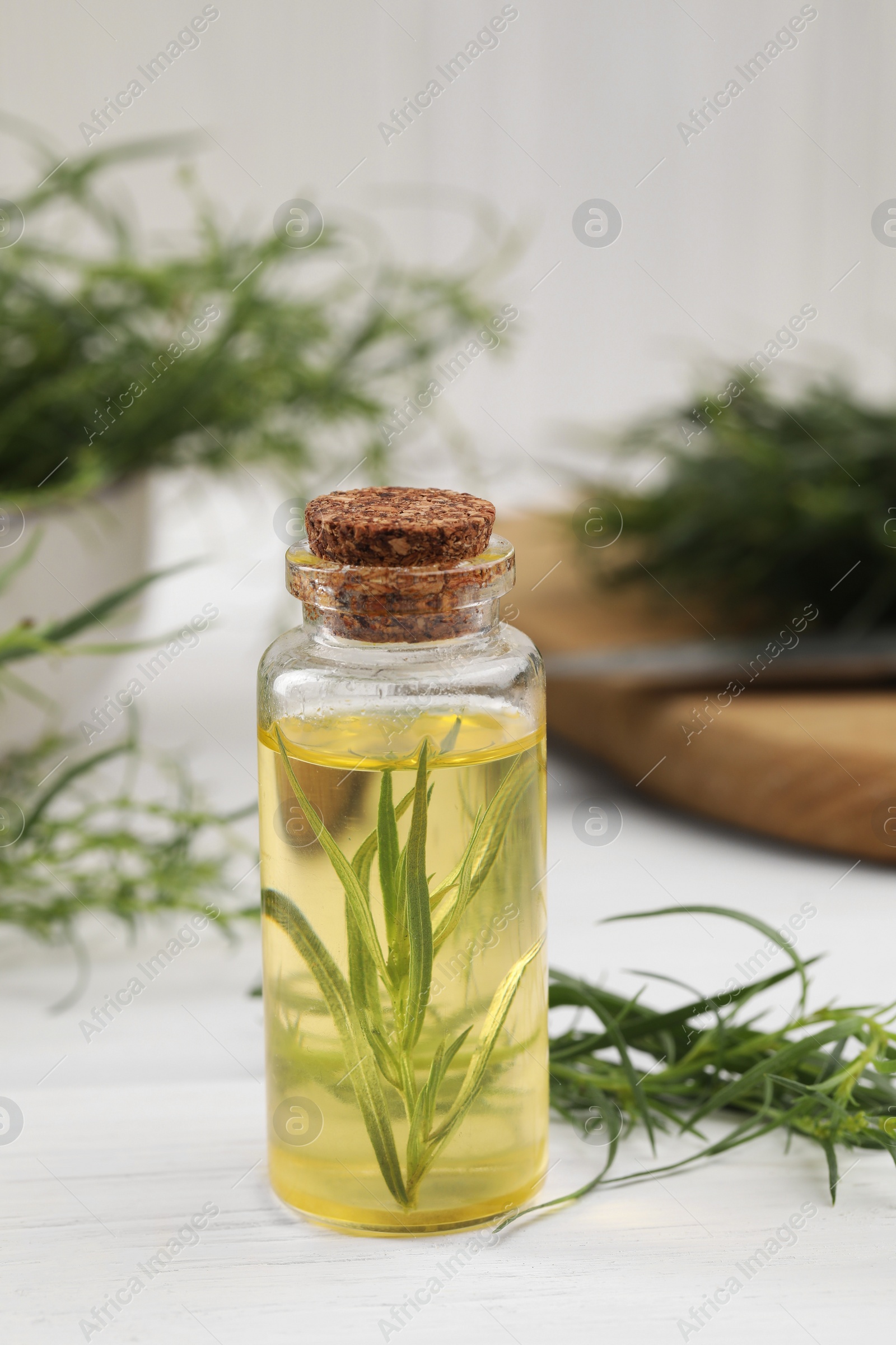 Photo of Bottle of essential oil and fresh tarragon leaves on white wooden table