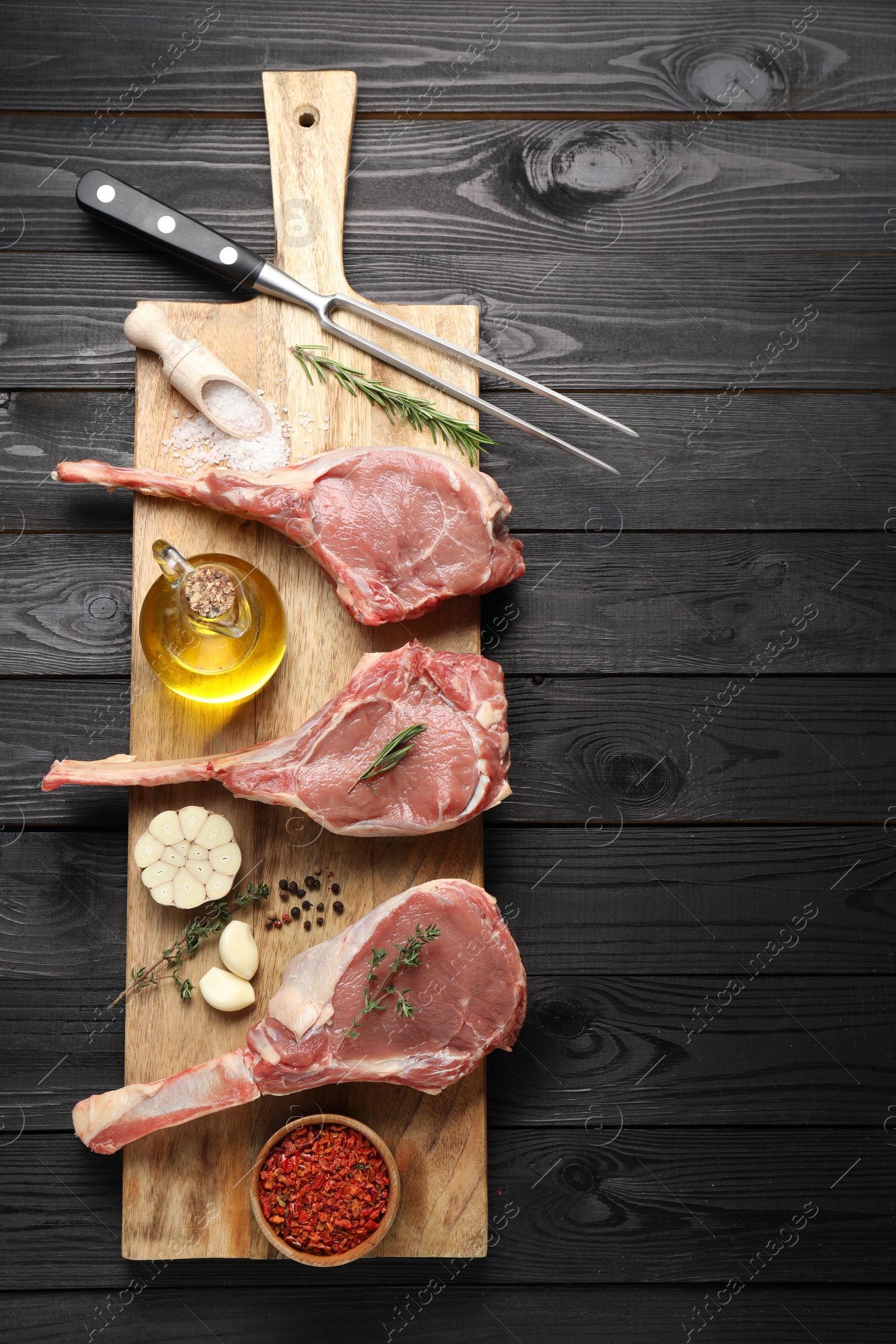 Photo of Fresh tomahawk beef cuts, spices and butcher fork on black wooden table, top view