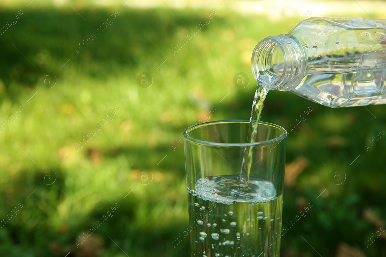 Photo of Pouring water from bottle into glass and blurred green grass on background. Space for text