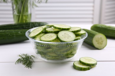 Photo of Cut cucumber in glass bowl, fresh vegetables and dill on white wooden table, closeup