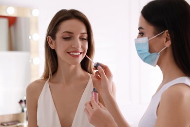 Photo of Makeup artist in protective mask working with woman indoors. Preventive measures during COVID-19 pandemic