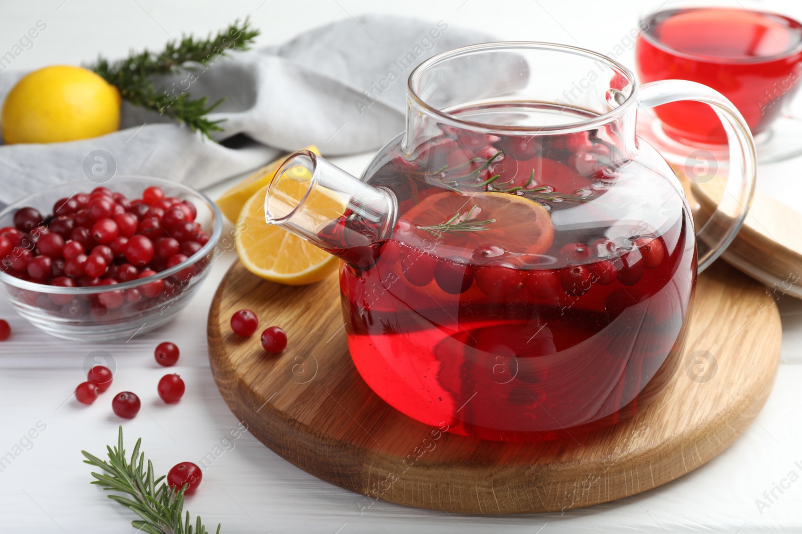 Photo of Tasty hot cranberry tea in teapot, rosemary and fresh berries on white wooden table