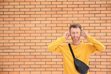 Young man listening to music with headphones against brick wall. Space for text