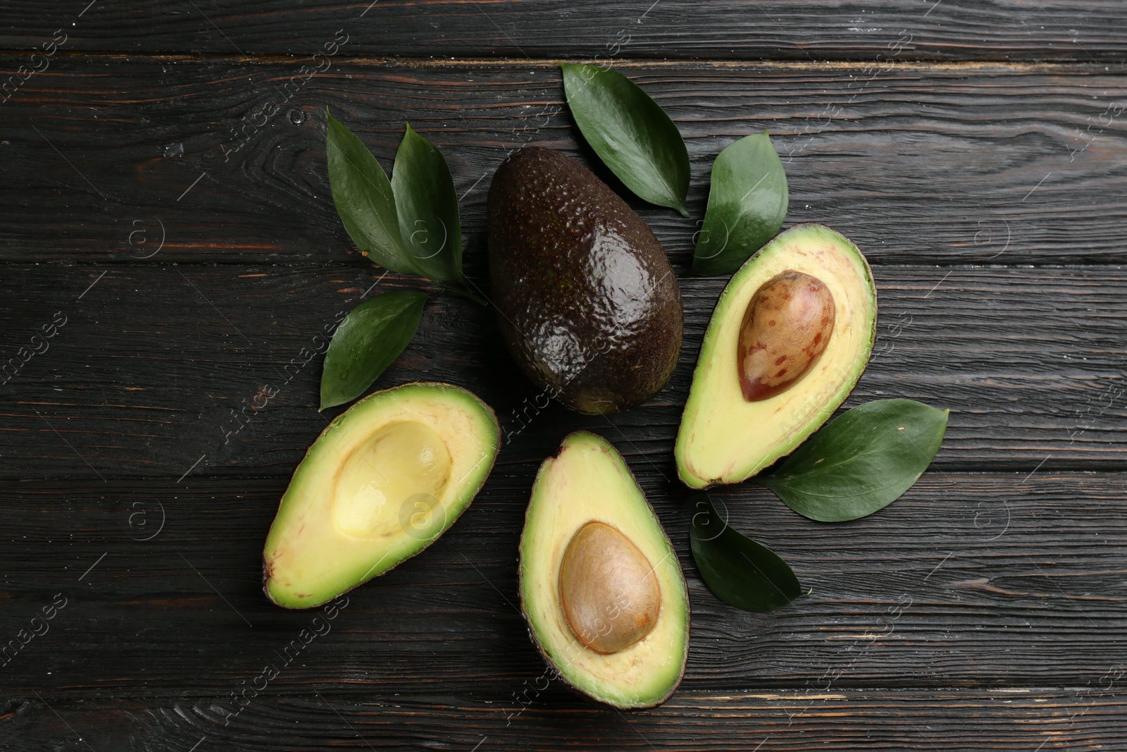 Photo of Whole and cut avocados with green leaves on dark wooden table, flat lay