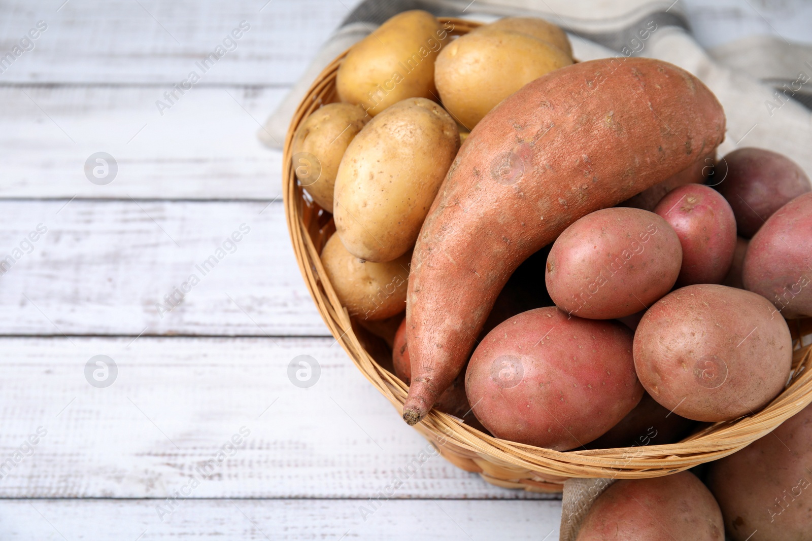 Photo of Different types of fresh potatoes in wicker basket on white wooden table, closeup. Space for text