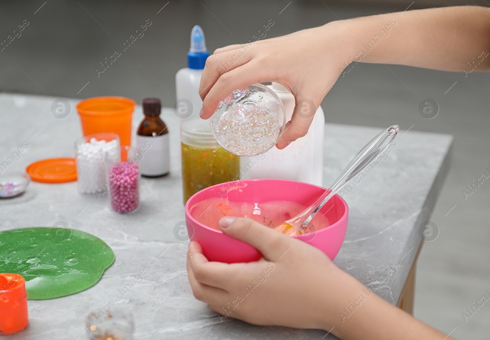 Photo of Little girl adding sparkles into homemade slime toy at table, closeup of hands