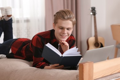 Online learning. Smiling teenage boy reading book near laptop on bed at home