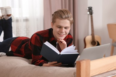 Photo of Online learning. Smiling teenage boy reading book near laptop on bed at home