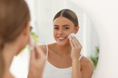 Photo of Beautiful woman removing makeup with cotton pad near mirror indoors