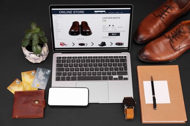 Photo of Online store website on laptop screen. Computer, smartphone, stationery, credit cards, men's shoes and accessories on black background, above view