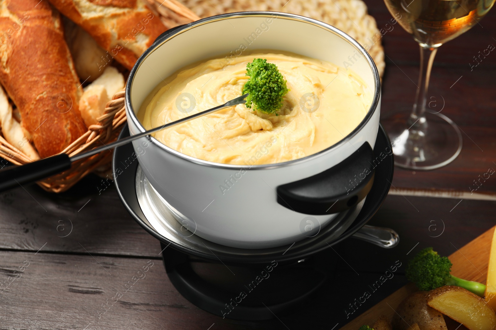 Photo of Fork with piece of broccoli, melted cheese in fondue pot and snacks on wooden table, closeup