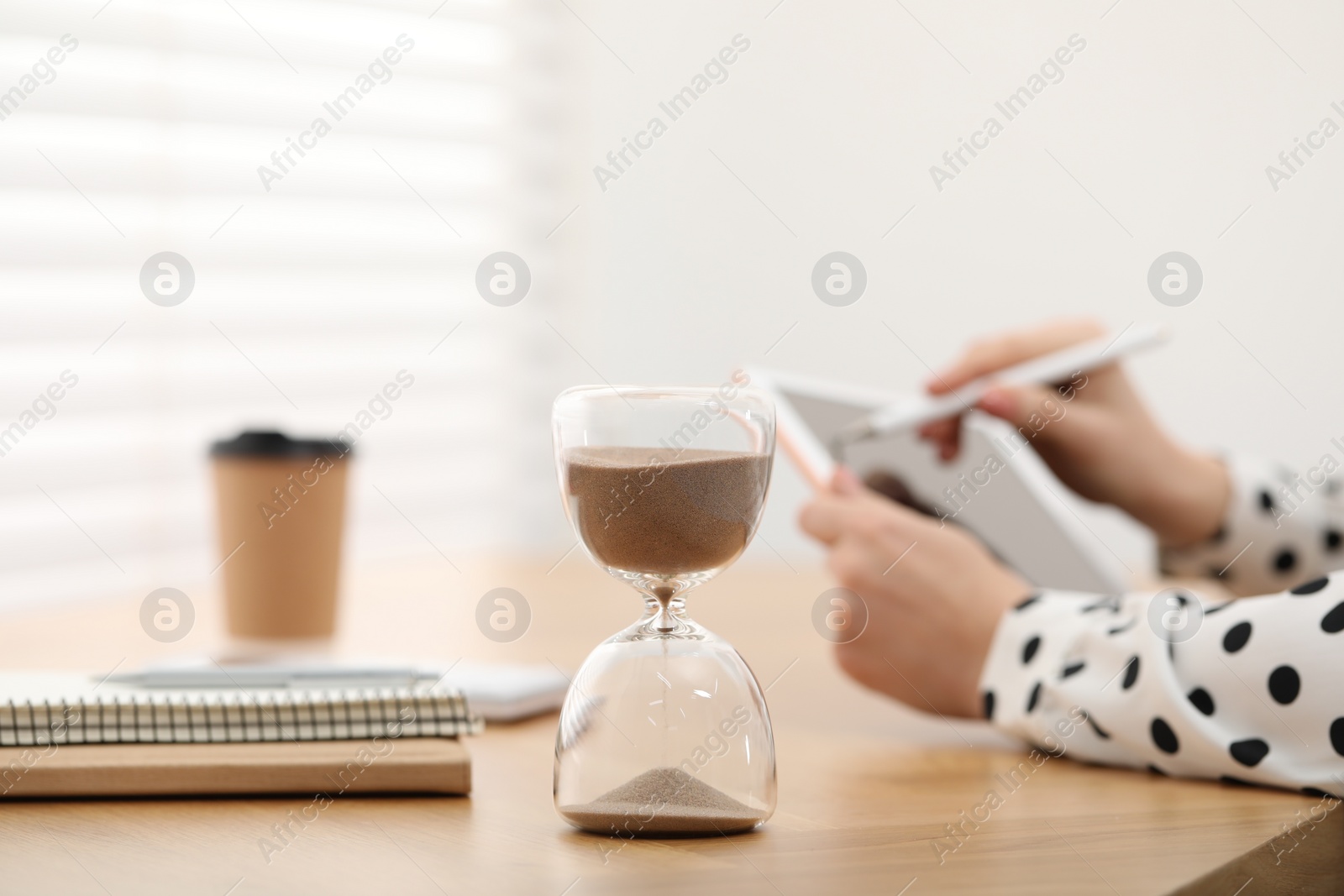 Photo of Hourglass with flowing sand on desk. Woman using tablet indoors, selective focus