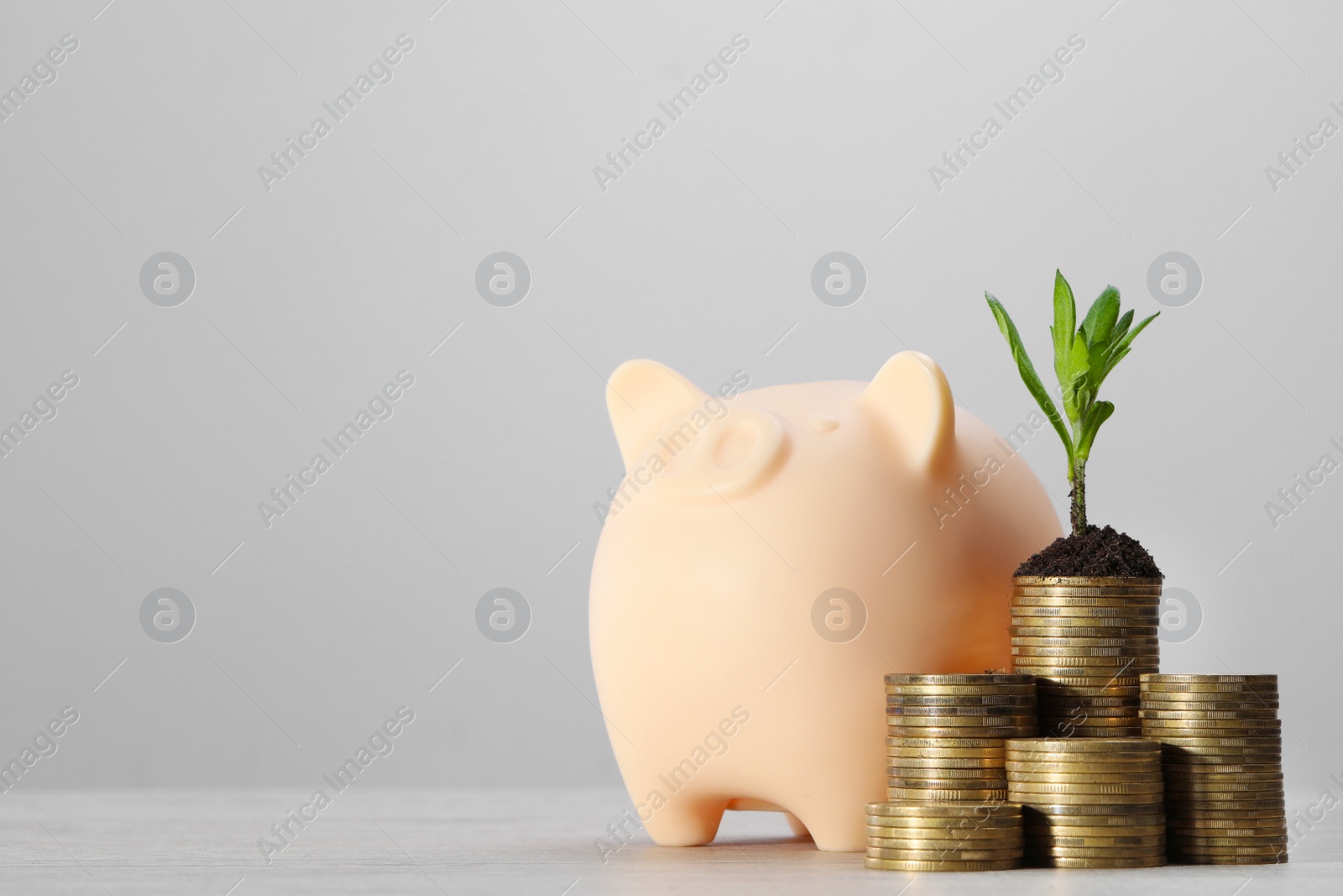 Photo of Stacks of coins with green seedling and piggy bank on white table against light grey background, space for text. Investment concept