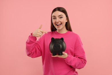 Photo of Happy woman pointing at piggy bank on pink background