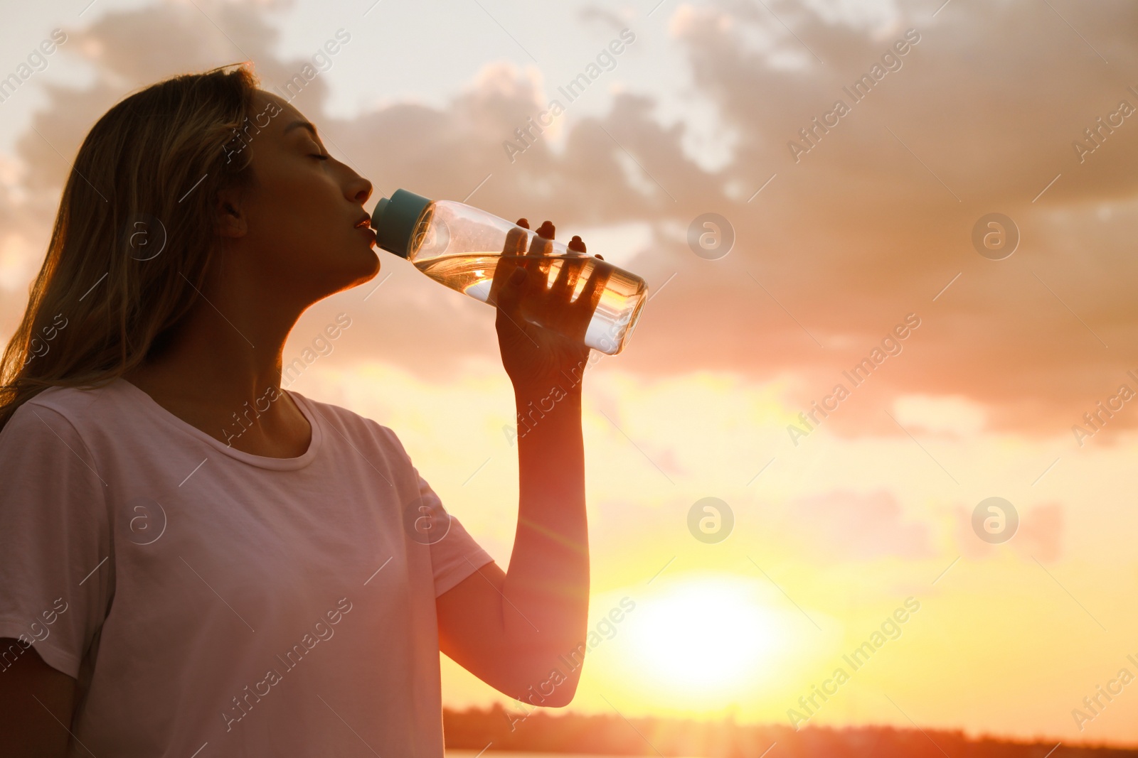 Photo of Young woman drinking water to prevent heat stroke outdoors at sunset, space for text