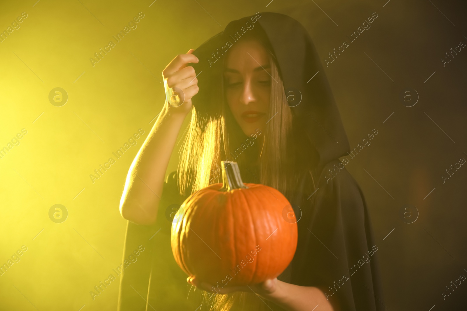 Photo of Young woman wearing witch costume with pumpkin in smoke cloud on dark background. Halloween party