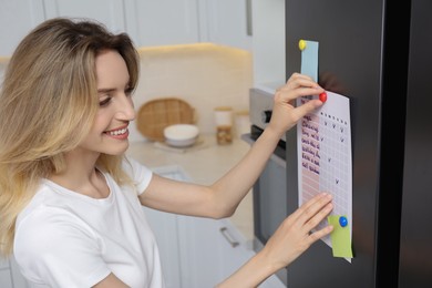 Photo of Young woman putting to do list on refrigerator door in kitchen