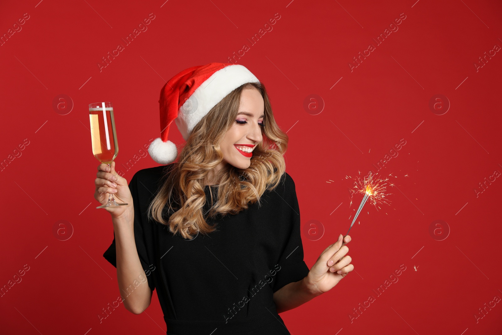 Photo of Happy young woman wearing Santa hat with glass of champagne and sparkler on red background. Christmas celebration