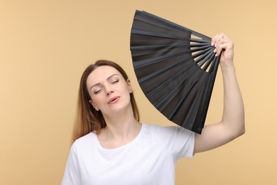 Beautiful woman waving black hand fan to cool herself on beige background