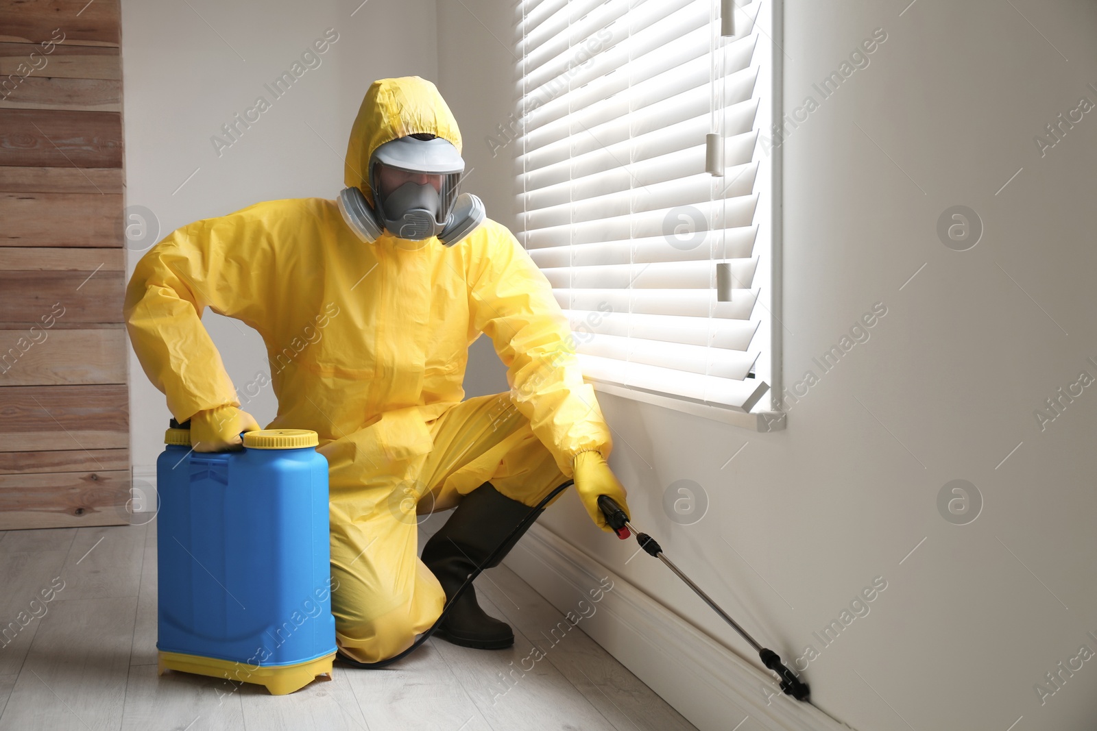 Photo of Pest control worker in protective suit spraying pesticide near window indoors